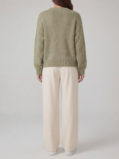 Person wearing a green knitted sweater and beige pants on a plain background